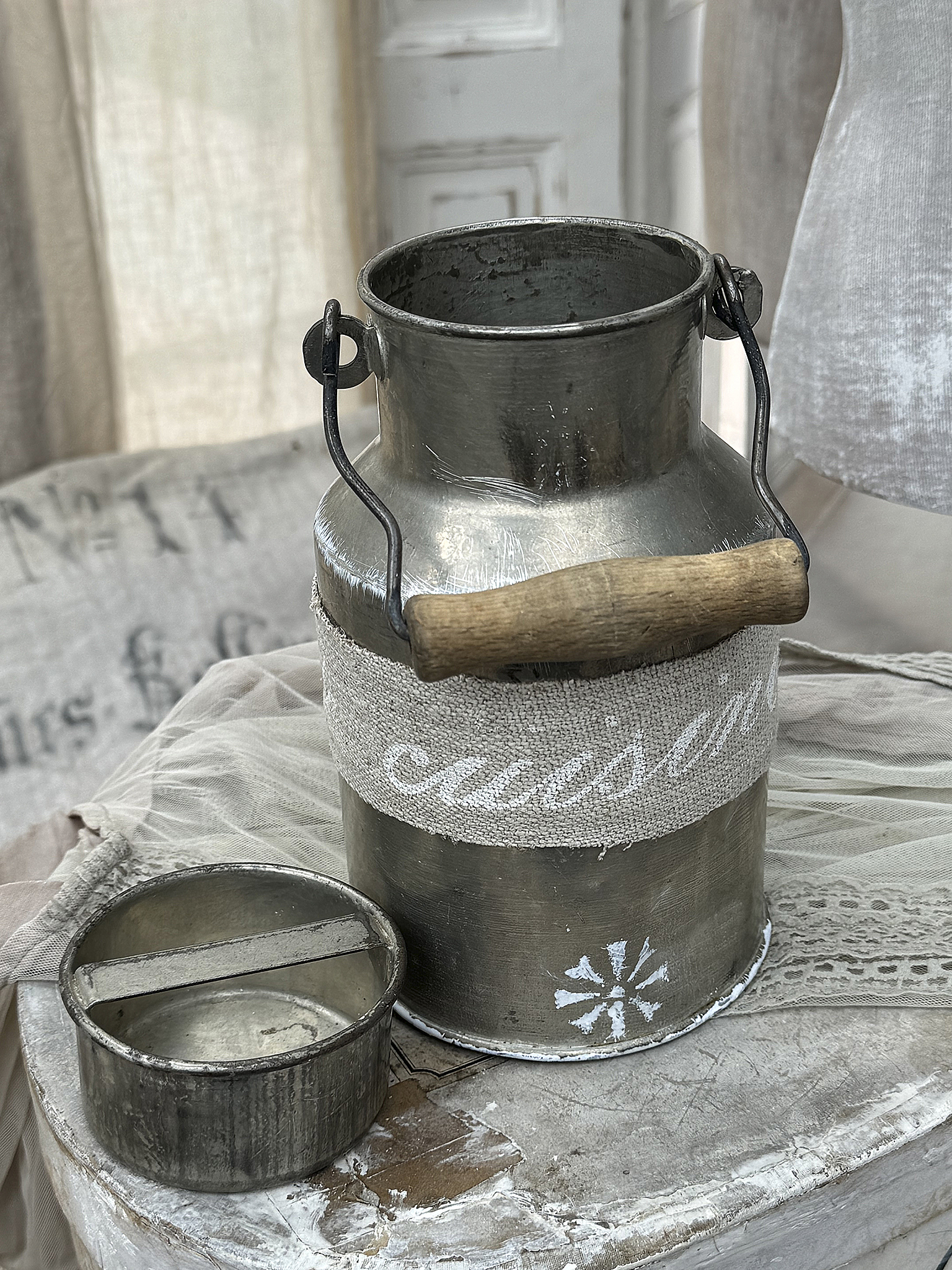 Dekorativer Milchkessel aus Metall mit Holzgriff und kleiner Metallschale, auf einem Tisch mit Vintage-Stoffen